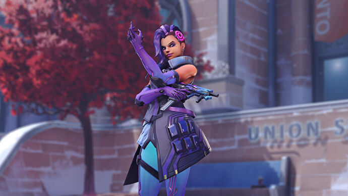 Sombra, a hero in Overwatch 2, poses outside a building in front of the camera, with a red-leaved tree behind her on the left.